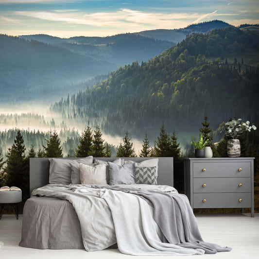 A nature-themed non-woven photo wallpaper featuring a mountain forest scenery, installed on a wall above a bed with gray bedding and pillows in a room with a drawer unit and a vase with flowers.