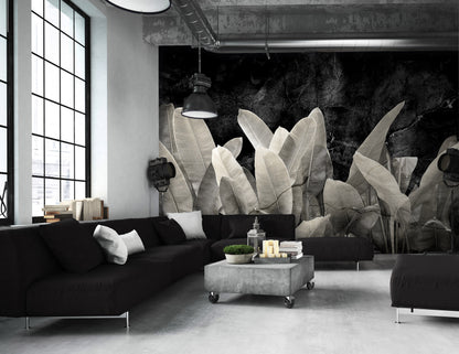 Modern living room with black sofa, gray ottoman, and large leafy plant wall mural.
